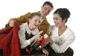 Two parents and their child are together against a white background. One parent is giving the child a wooden nutcracker, and the other parent is putting a red blanket around the child.